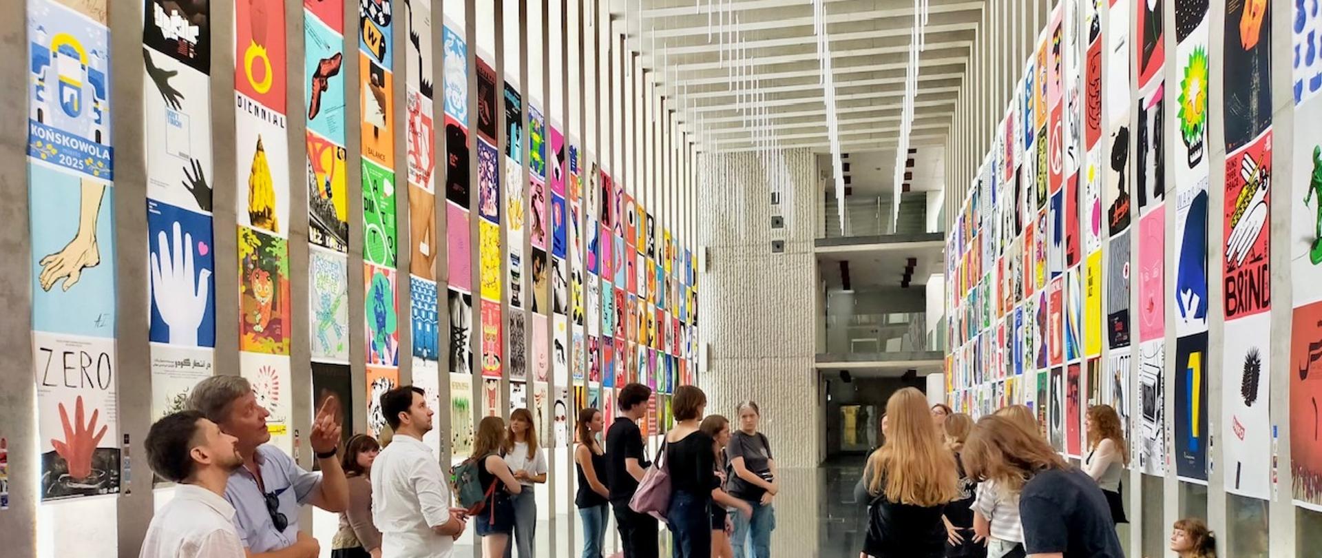 Na zdjęciu widać przestronną, nowoczesną salę wystawową z wysokim sufitem i dużymi oknami po lewej stronie. Na obu długich ścianach zawieszono setki kolorowych plakatów w równych rzędach, tworzących efektowną galerię. Każdy plakat przedstawia inny motyw graficzny – od abstrakcyjnych kształtów, przez rysunki i symbole, po napisy. W centralnej części przestrzeni znajduje się grupa osób – młodzi ludzie i dorośli – którzy oglądają prace, rozmawiają ze sobą, wskazują na plakaty, a część z nich robi zdjęcia. Atmosfera jest swobodna i skupiona, typowa dla wystawy sztuki współczesnej.