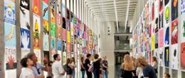 Na zdjęciu widać przestronną, nowoczesną salę wystawową z wysokim sufitem i dużymi oknami po lewej stronie. Na obu długich ścianach zawieszono setki kolorowych plakatów w równych rzędach, tworzących efektowną galerię. Każdy plakat przedstawia inny motyw graficzny – od abstrakcyjnych kształtów, przez rysunki i symbole, po napisy. W centralnej części przestrzeni znajduje się grupa osób – młodzi ludzie i dorośli – którzy oglądają prace, rozmawiają ze sobą, wskazują na plakaty, a część z nich robi zdjęcia. Atmosfera jest swobodna i skupiona, typowa dla wystawy sztuki współczesnej.