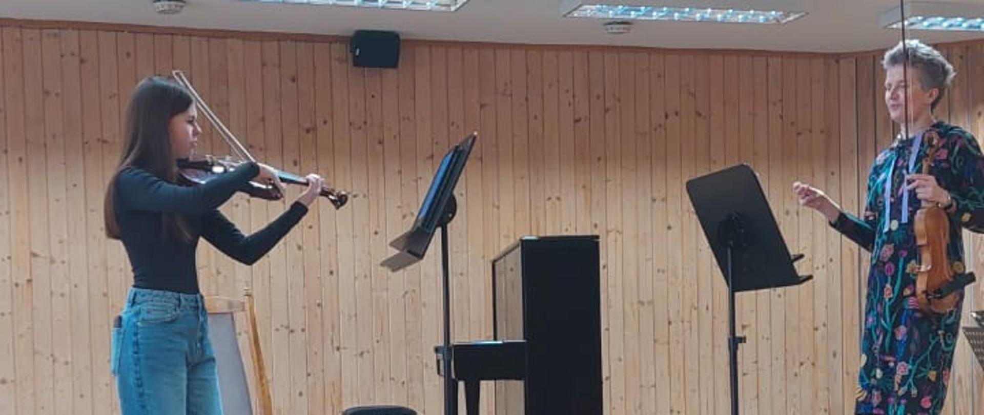 Nastolatka gra na skrzypcach, obok stoi kobieta.