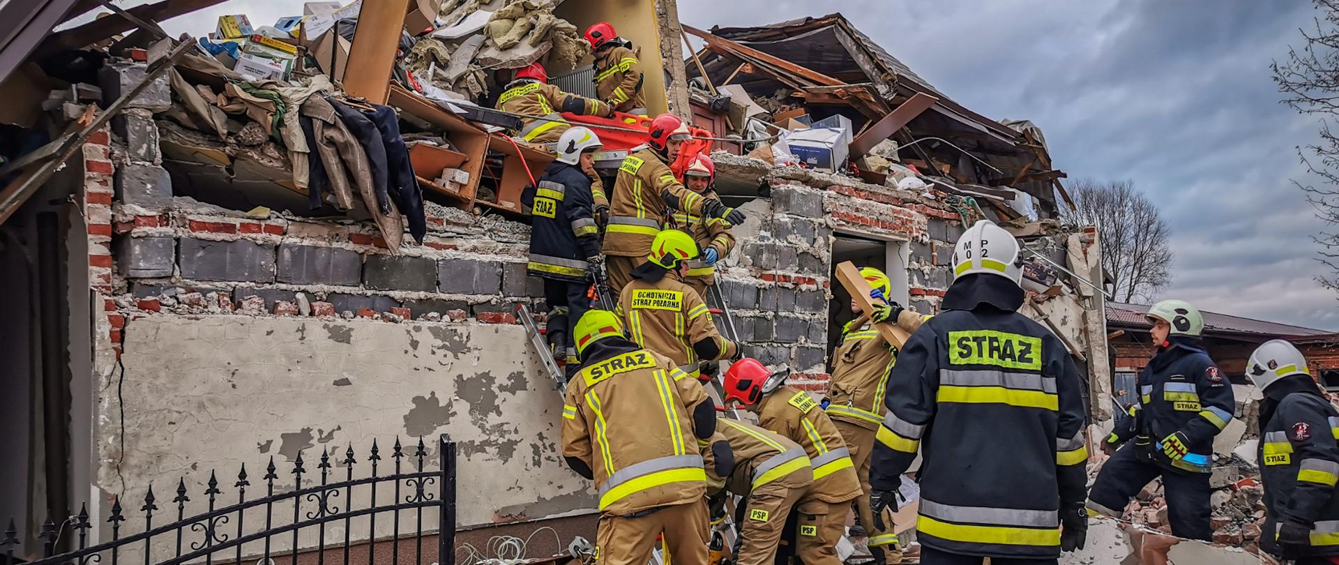 Na zdjęciu strażacy w akcji w ubraniach koloru musztardowego na drabinach przystawnych na tle budynku zawalonego w wyniku wybuchu gazu, wokół porozrzucane elementy budynku, dach wyrzucony poza budynek.