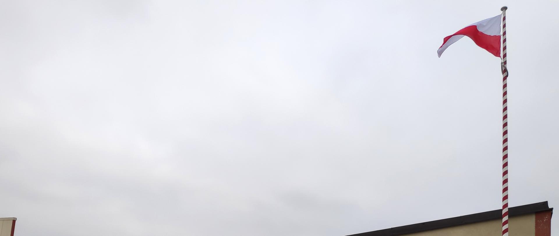 foto przedstawia strażaków podczas uroczystego apelu z okazji Narodowego święta Niepodległości, strażacy w umundurowaniu, hełmach i maseczkach, flaga Polski powiewa na wietrze