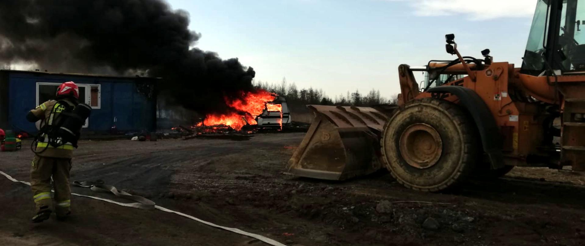 Zdjęcie przedstawia na środku palącą się przyczepę, a nad przyczepą widać kłęby czarnego dymu.Z lewej strony biegnie strażak w ubraniu specjalnym oraz aparacie ochrony układu oddechowego. Z prawej strony widać fragment spycharki.