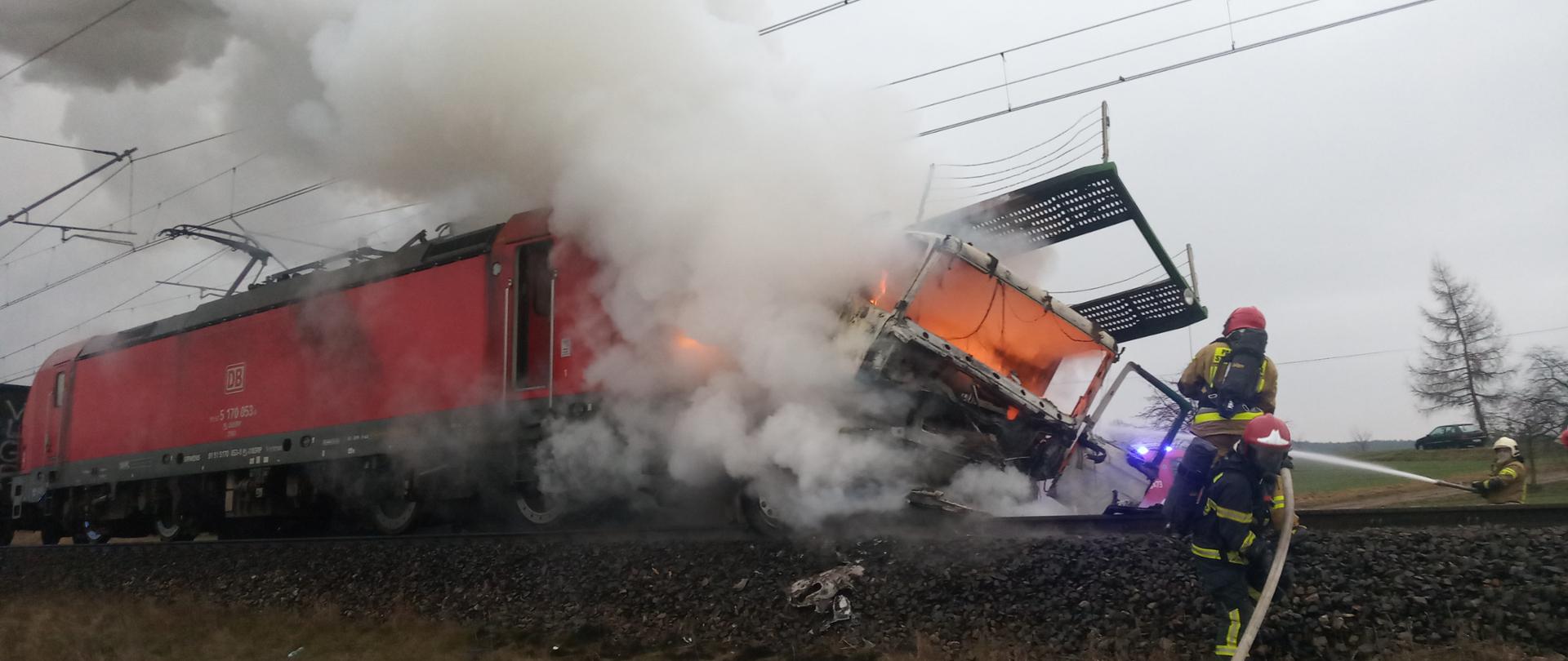 Na zdjęciu widać płonący samochód ciężarowy po zderzeniu z lokomotywą. Jest duże zadymienie a strażacy zabezpieczeni w aparaty ochrony dróg oddechowych gaszą pożar