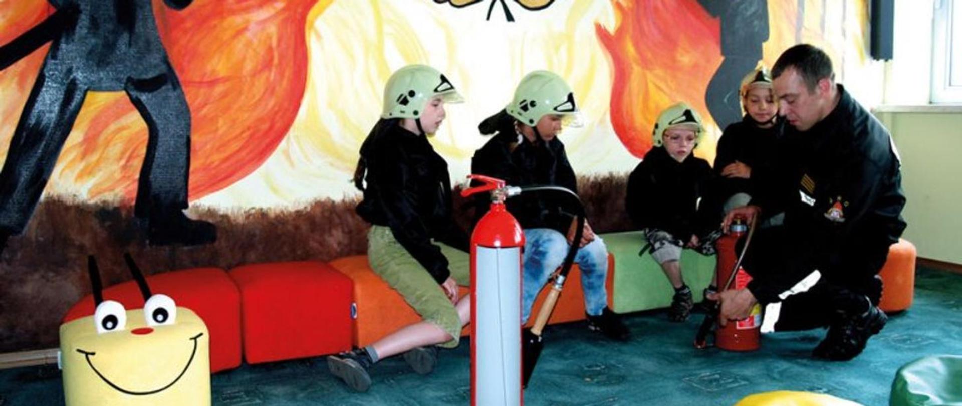 Zdjęcie przedstawia strażaka w ubraniu koszarowym demonstrującego czwórce dzieci zasadę działania gaśnicy. Dzieci mają na głowie plastikowe hełmy i siedzą na kanapie która przypomina stonogę. Na ścianie za nimi widać grafikę przedstawiającą strażaka gaszącego pożar