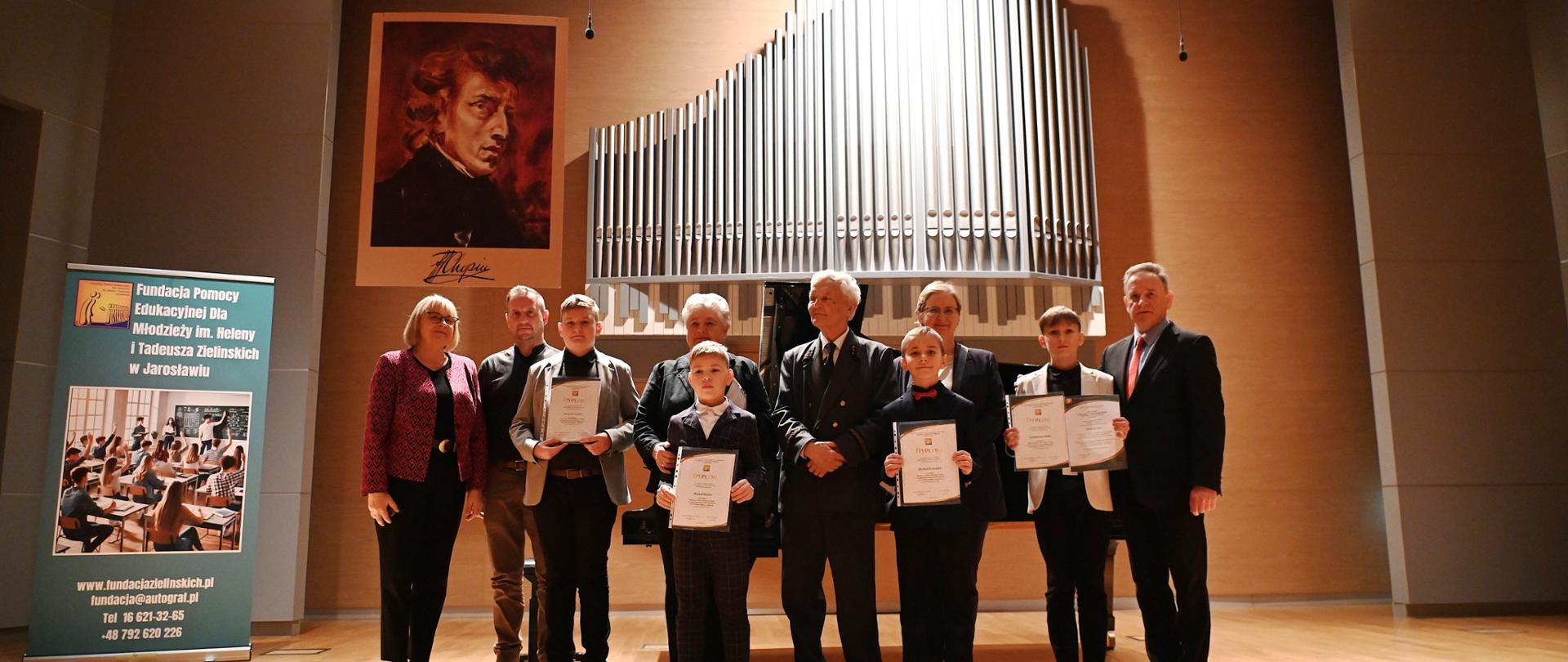 Fotografia przedstawiająca grupę osób stojących na drewnianej scenie sali koncertowej. W grupie znajdują się dorośli (organizatorzy i nauczyciele) oraz uczniowie w odświętnych strojach, trzymający w rękach dyplomy. W tle widać piszczałki organowe oraz duży portret Fryderyka Chopina wiszący na ścianie. Po lewej stronie stoi roll-up z logo Fundacji Pomocy Edukacyjnej dla Młodzieży im. Heleny i Tadeusza Zielińskich w Jarosławiu.