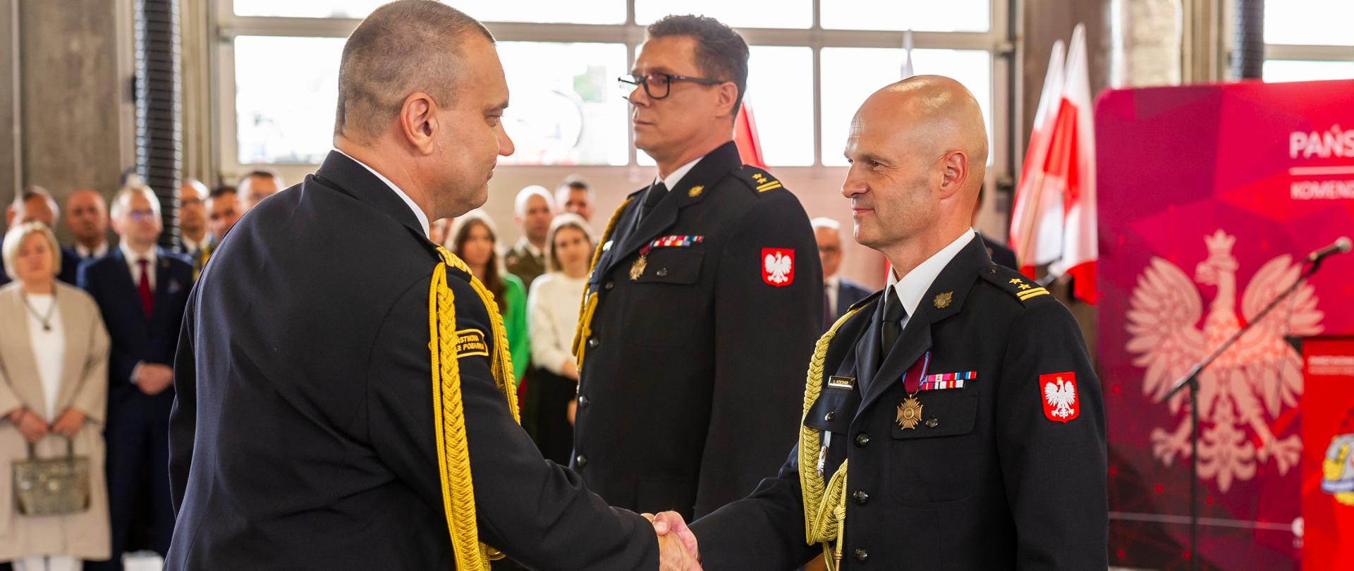 Komendant wojewódzki ściska rękę ustępującemu komendantowi. Obok znajdują się nowy komendant miejski, w tle sylwetki zgromadzonych gości.