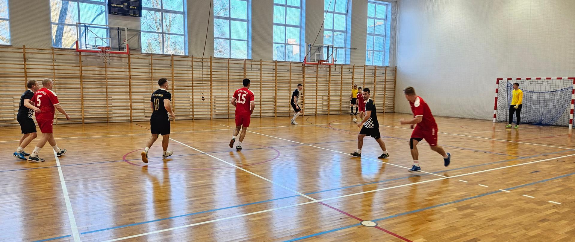 Dynamiczna akcja na połowie boiska. Zawodnicy w czerwonych strojach (numer 15 i 5) oraz zawodnicy w czarno-białych koszulkach poruszają się po parkiecie. W tle widać bramkarza w żółtej bluzie stojącego w bramce.