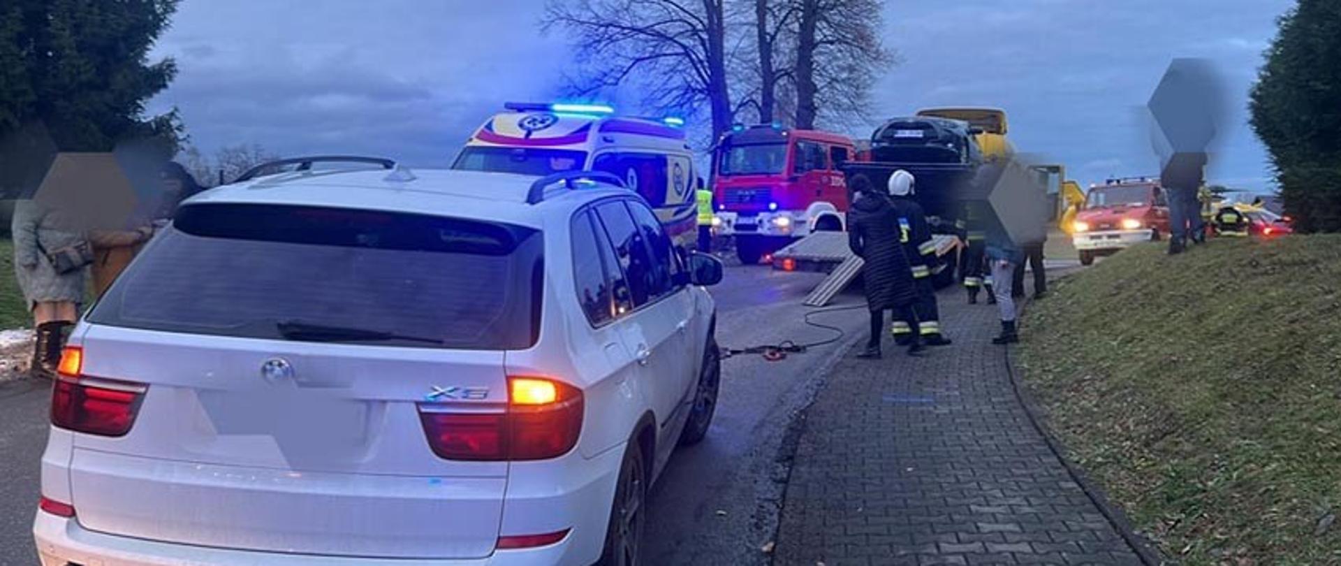 Widok z tyłu. Pora wieczorna. Na pierwszym planie pojazd, który przygniótł mężczyznę. W oddali widać strażaków oraz pojazdy służb ratunkowych.