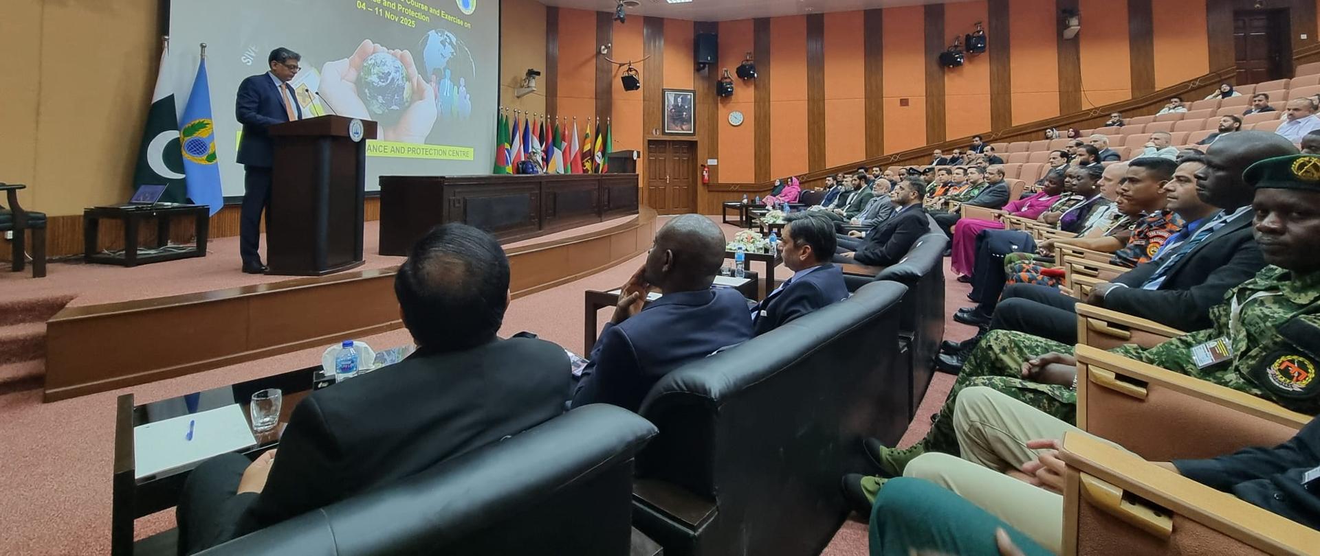 Aula wypełniona siedzącymi w niej uczestnikami konferencji, po lewej na podwyższeniu mównica przy której stoi prelegent, w tle ekran multimedialny z wyświetlonym slajdem prezentacji