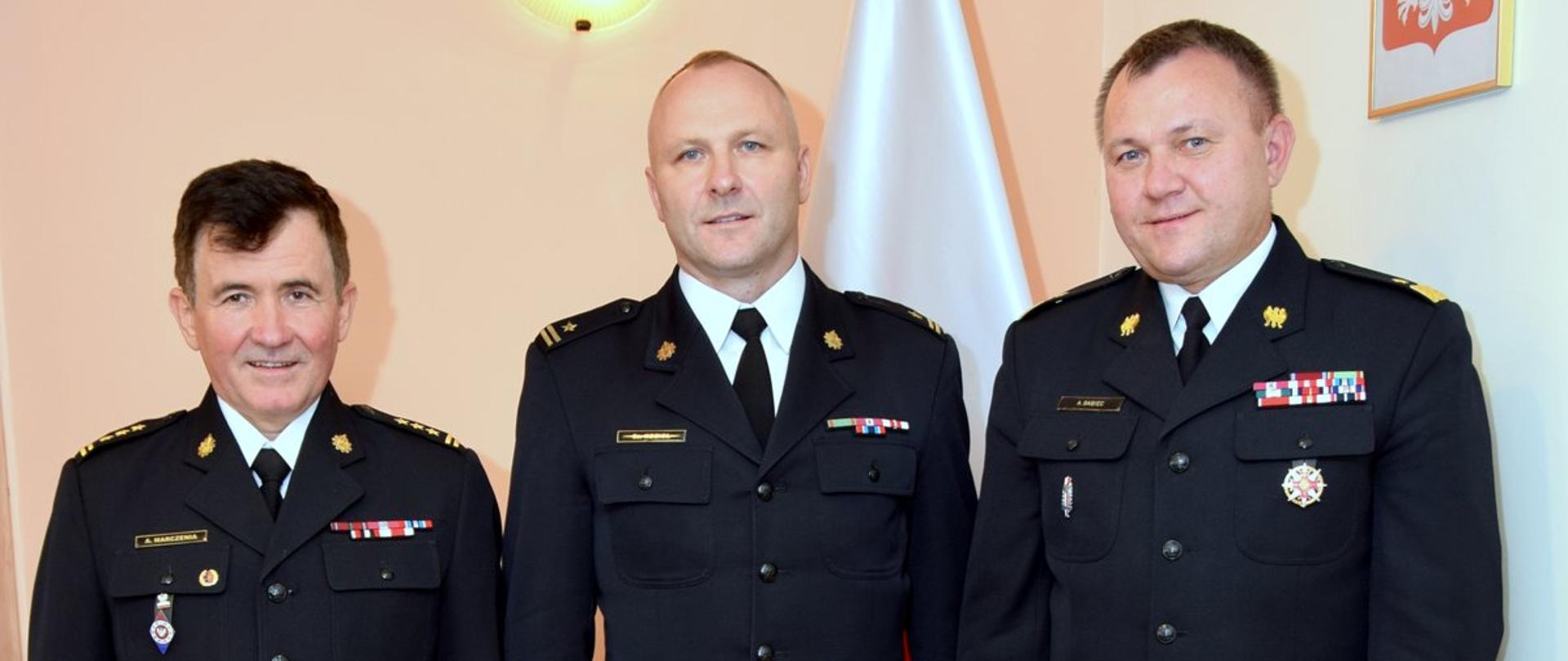 Zdjęcie zrobione w pomieszczeniu. W szeregu, w umundurowaniu wyjściowym stoją trzej funkcjonariusze Państwowej Straży Pożarnej. Od lewej strony: funkcjonariusz w stopniu starszego brygadiera, młodszego brygadiera i generała.
