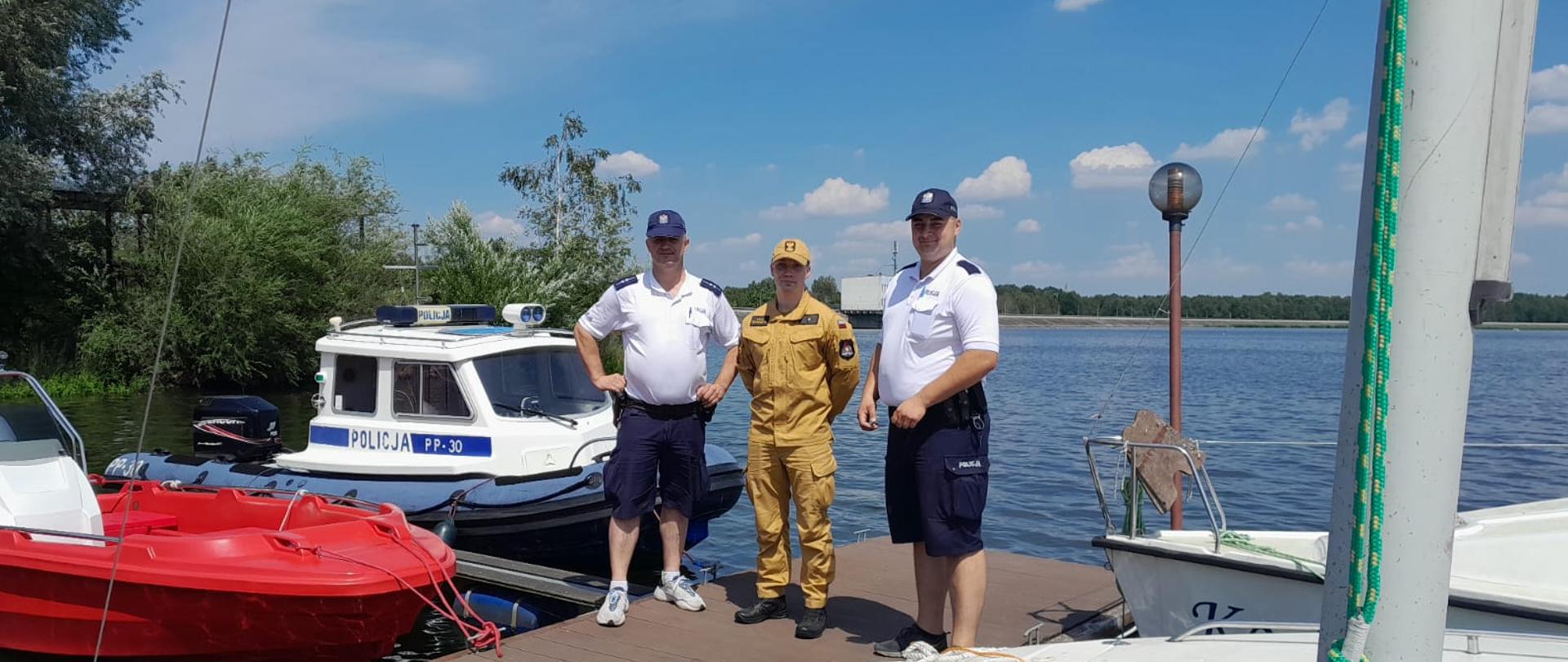 Funkcjonariusz Państwowej Straży Pożarnej i dwóch funkcjonariuszy Policji podczas wspólnych działań na rzecz bezpieczeństwa nad wodą. W tle policyjna łódź oraz zbiornik wodny Poraj.