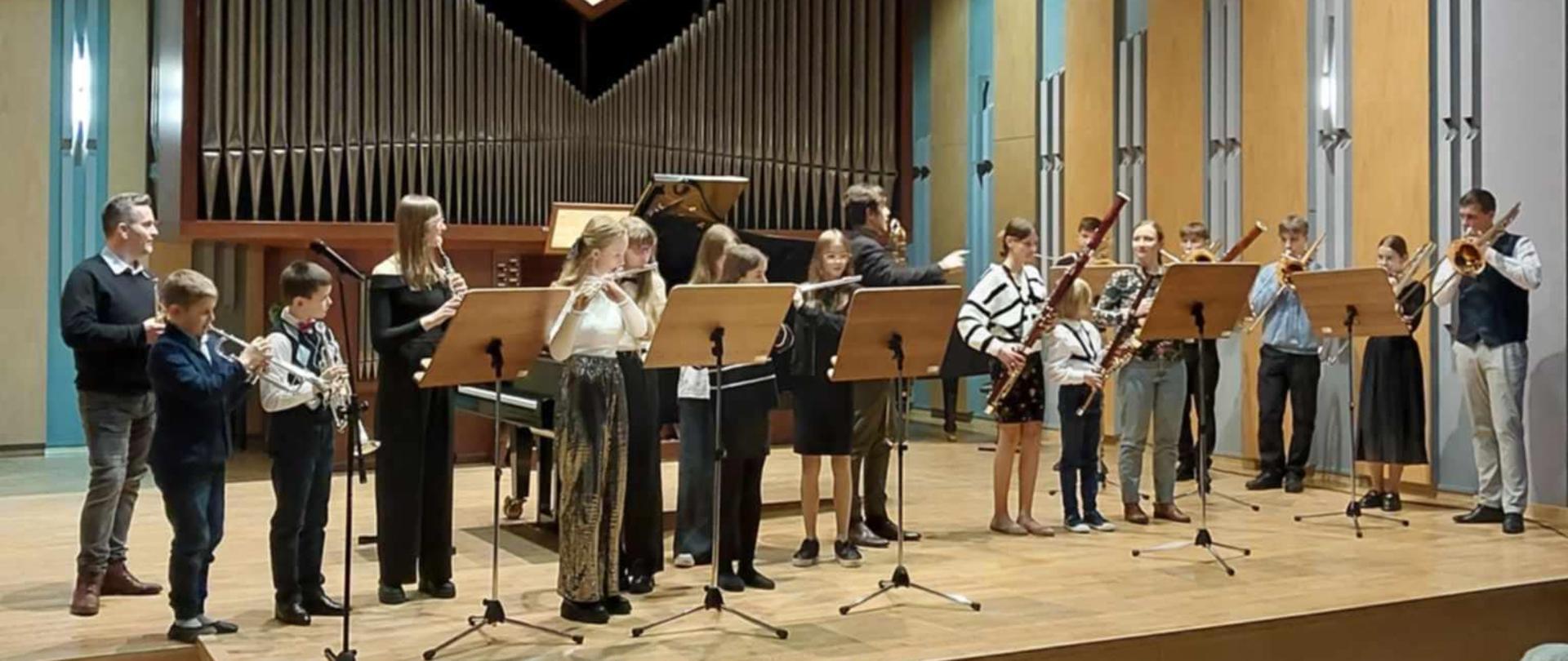 Wszyscy uczestnicy koncertu stoją na scenie sali koncertowej trzymając w dłoni swoje instrumenty. Przed nimi pulpity. Za nimi organy, Na suficie zapalone lampy, na ścianach kinkiety. Na pierwszym planie widownia oraz kilka pustych foteli