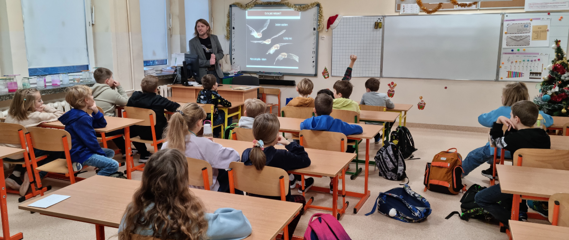 Sala w szkole. Dzieci siedzą przy ławkach. Pracownik Regionalnej Dyrekcji Ochrony Środowiska w Olsztynie opowiada dzieciom o nietoperzach.