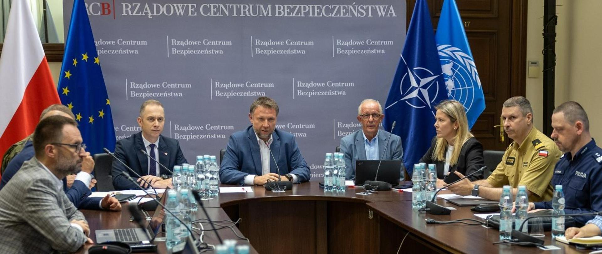 Grupa osób na czele z ministrem Kierwińskim siedzi przy stole. Na stole woda mineralna, komputery i mikrofony. W tle baner Rządowego Centrum Zarządzania Kryzysowego oraz flagi: polska, unijna, NATO i ONZ. 