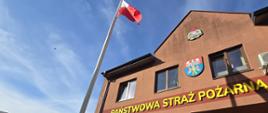 Zdjęcie przedstawia powiewającą na maszcie flagę biało-czerwoną. W tle budynek z napisem Państwowa Straż Pożarna oraz godła powiatu wołomińskiego oraz PSP.