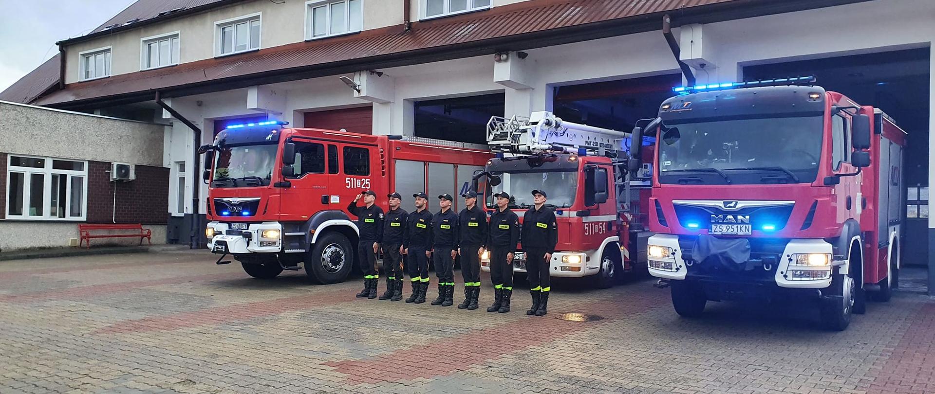 Trzy samochody pożarnicze na placu komendy z włączonymi sygnałami świetlno- dźwiękowymi oraz siedmiu strażaków oddających hłod zmarłemu na służbie koledze.