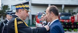 Na zdjęciu widoczni Małopolski Komendant Wojewódzki PSP wręczający osobie cywilnej w granatowym garniturze odznaczenie resortowe. Komendant w galowym granatowym mundurze i w czapce rogatywce. 