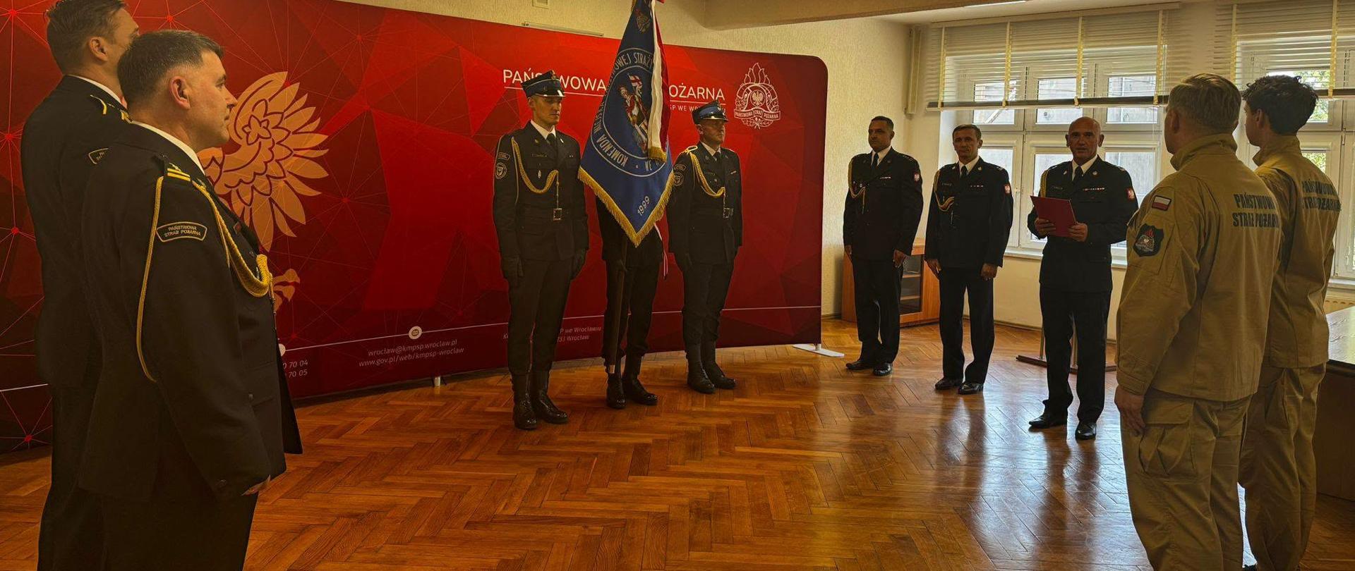 Uroczystość w siedzibie Państwowej Straży Pożarnej we Wrocławiu. Funkcjonariusze PSP w umundurowaniu galowym oraz strażacy w ubraniach specjalnych stoją podczas ceremonii przed pocztem sztandarowym