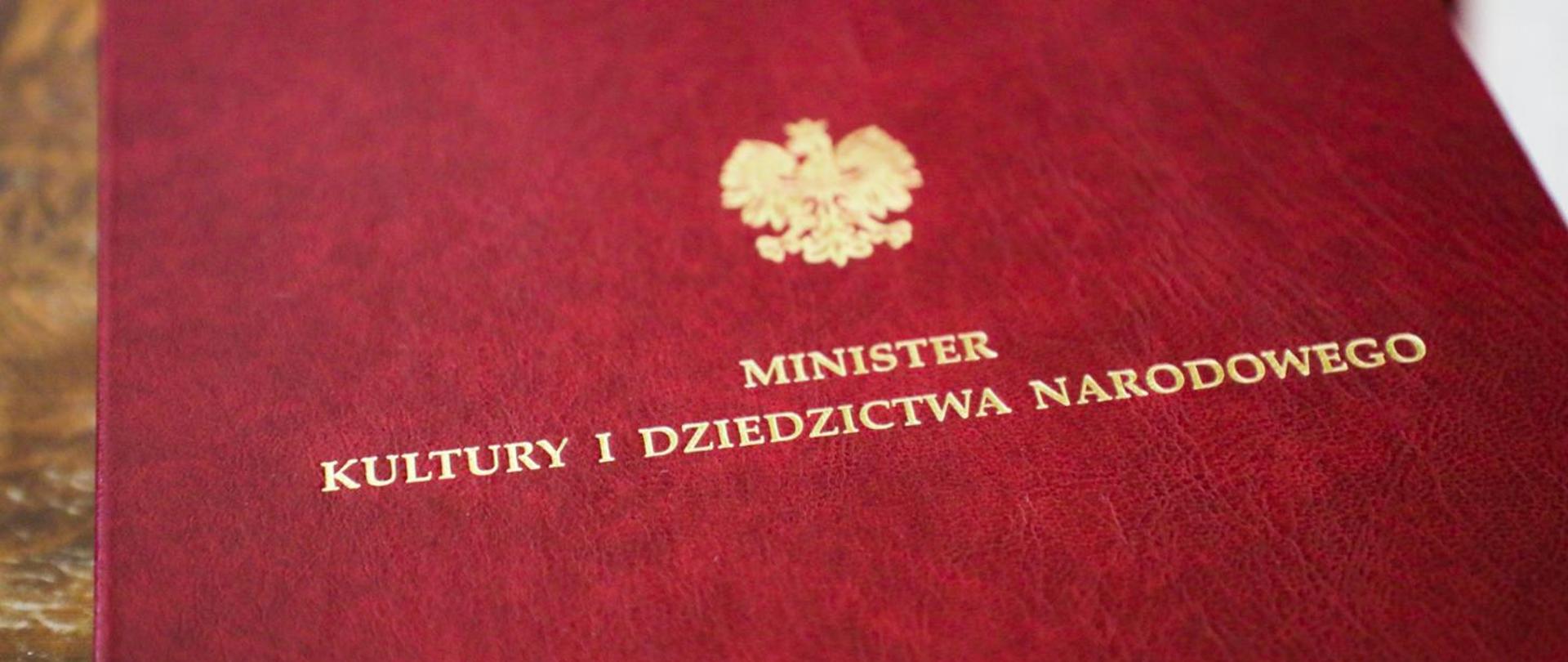 grafika przedstawia fragment czerwonej teczki ze złotym orłem po środku oraz napisem z napisem minister kultury i dziedzictwa narodowego