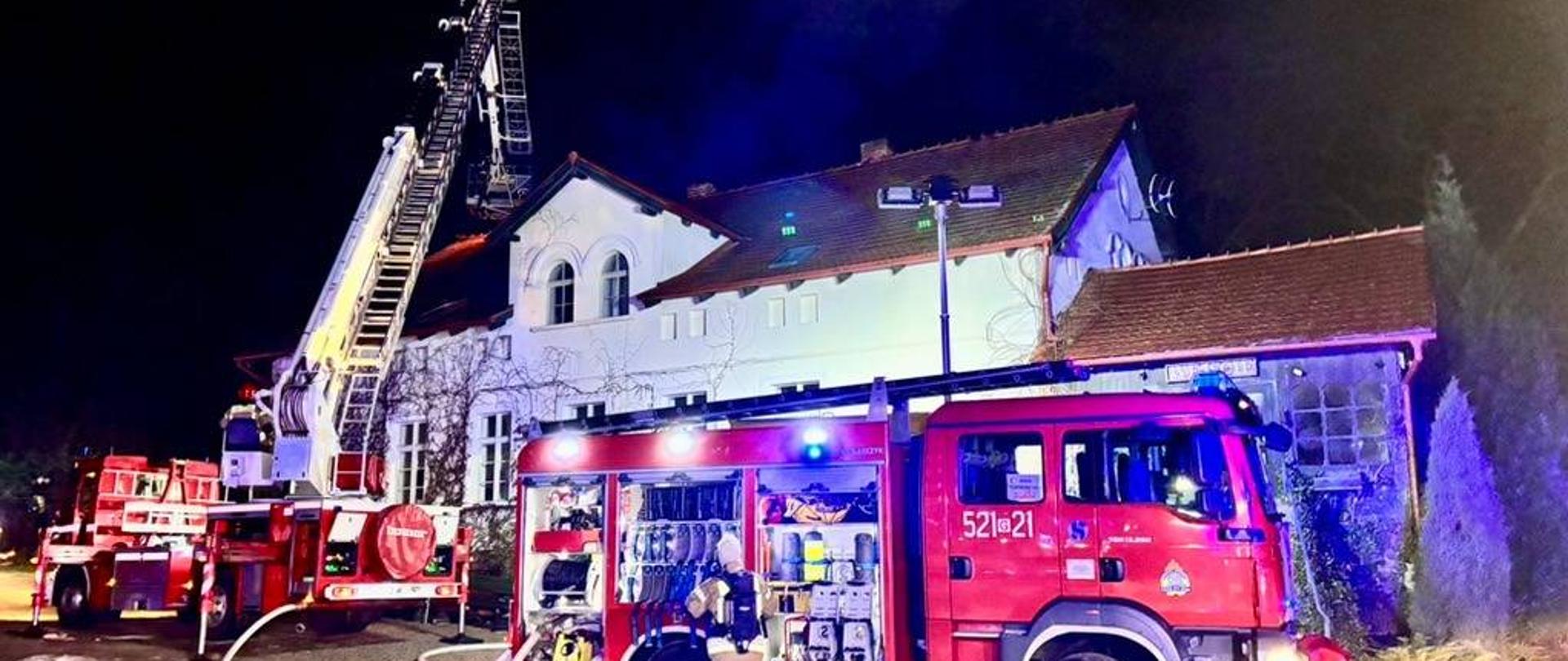 Zdjęcie przedstawia samochody pożarnicze- gaśniczy i drabinę przed budynkiem objętym pożarem. 