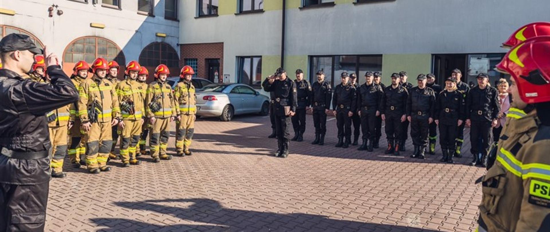 Uroczysta zmiana służby na placu wewnętrznym Komendy Powiatowej PSP w Tarnowskich Górach. Na zbiórce obecni strażacy w ubraniach specjalnych koloru piaskowego oraz kadra dowódcza. 