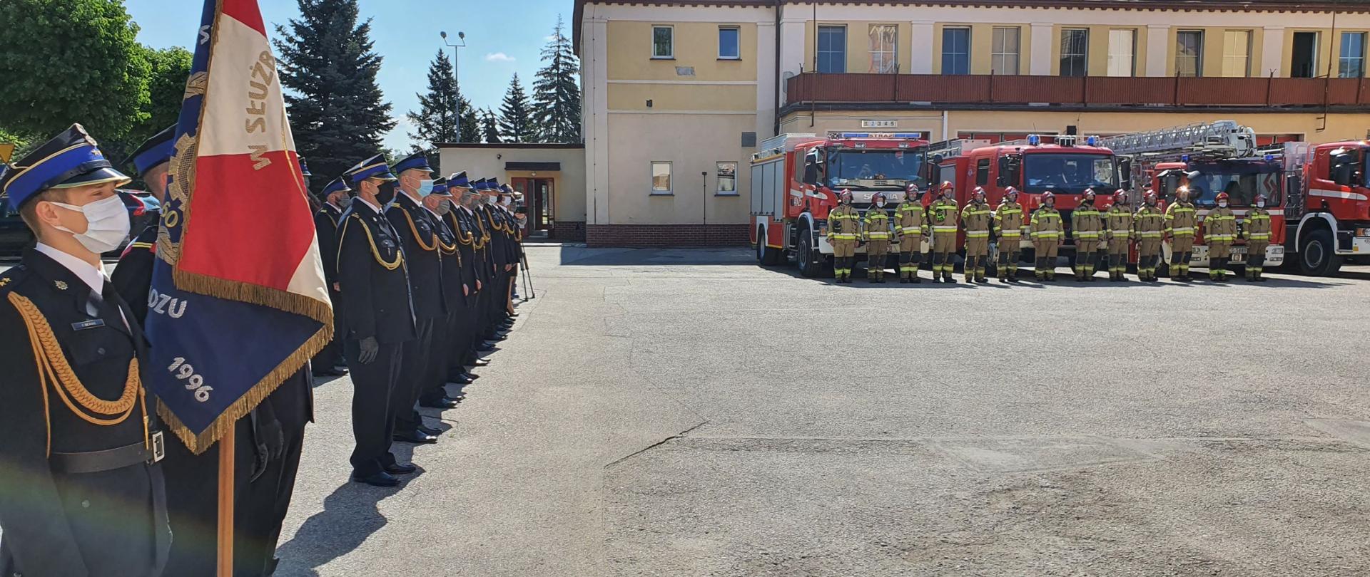 Zdjęcie przedstawia strażaków podczas uroczystej zbiórki. Po lewej funkcjonariusze w mundurach galowych na wprost przed czerwonymi samochodami gaśniczymi strażacy w piaskowych mundurach specjalnych. W tle budynek strażnicy.