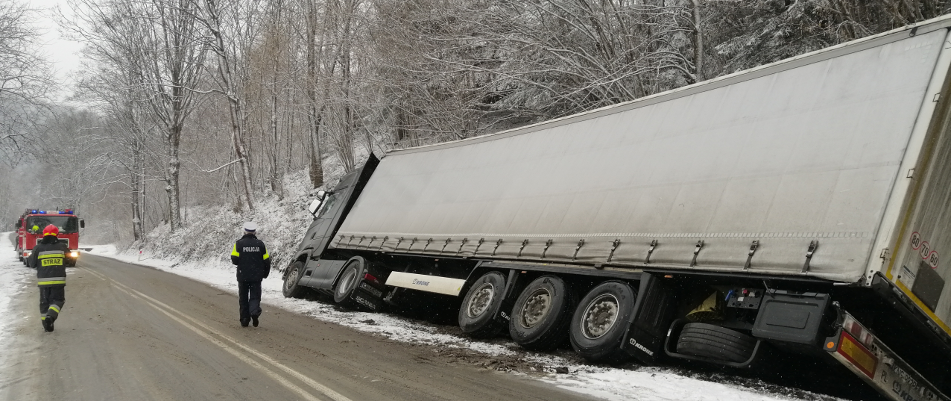 Samochód ciężarowy w rowie. Na drodze policjant oraz strażak. W tle samochód pożarniczy.