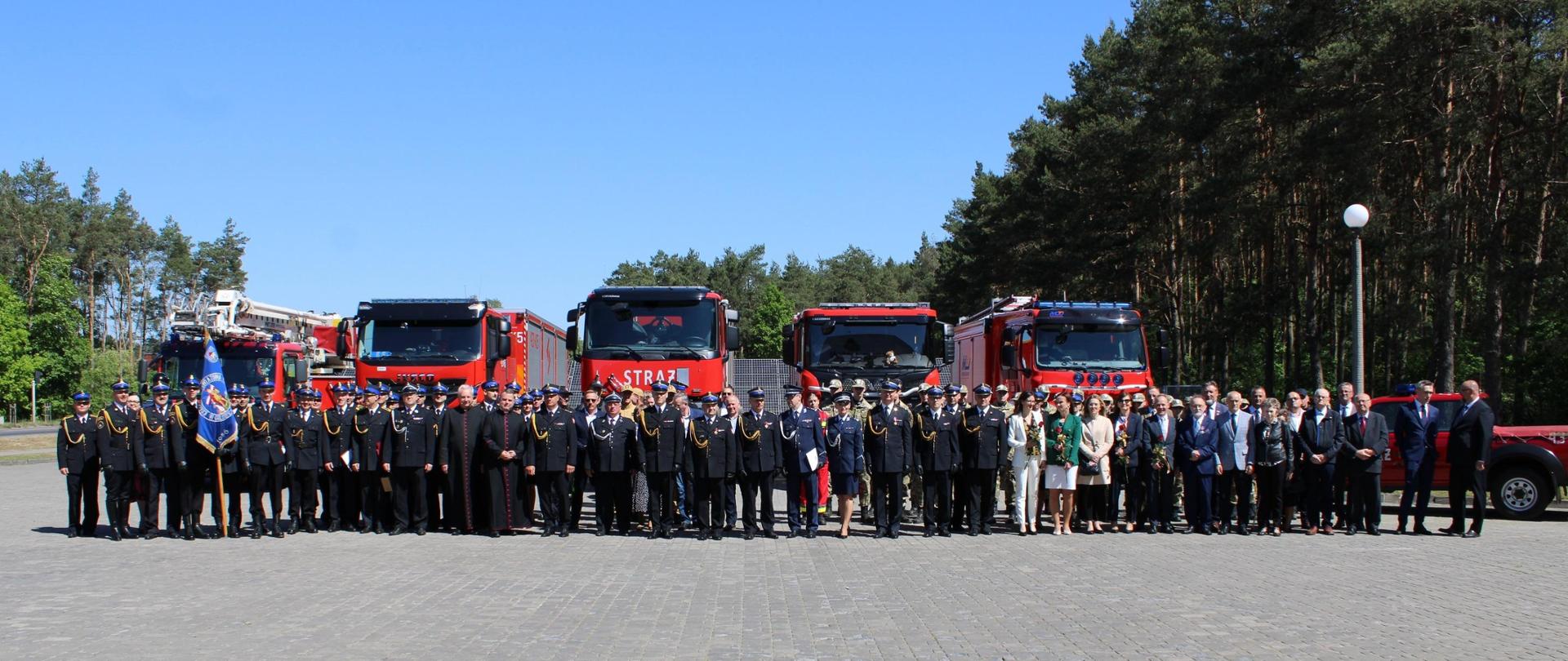 Zebrani goście oraz funkcjonariusze uczestniczący w zbiórce stają w szeregu, w tle widać wozy ratowniczo-gaśnicze.