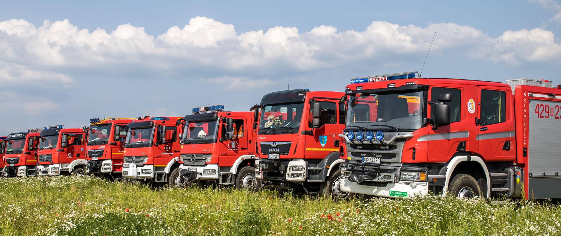 Zdjęcie przedstawia ustawione jeden obok drugiego, przodem do fotografującego samochody pożarnicze koloru czerwonego. Z przodu łąka kwiecista. 