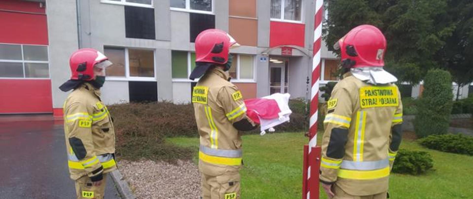Trzech strażaków przed budynkiem PSP , jeden (w środku) trzyma przed sobą na rękach złożoną Flagę Polską, która za chwilę zostanie uroczyście podniesiona na maszt