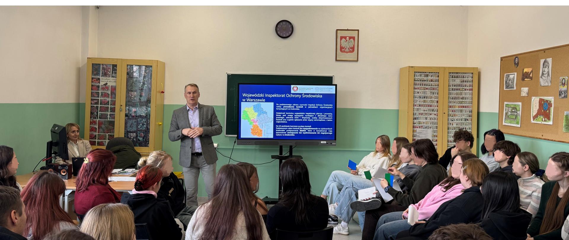 Inspektor Wojewódzkiego Inspektoratu Ochrony Środowiska przedstawia w szkolnej klasie prezentację dotyczącą Wojewódzkiego Inspektoratu Ochrony Środowiska. Przed nim siedzi grupa uczniów trzymając kolorowe kartki. W tle widać szkolne gabloty, tablicę i godło Polski na ścianie.