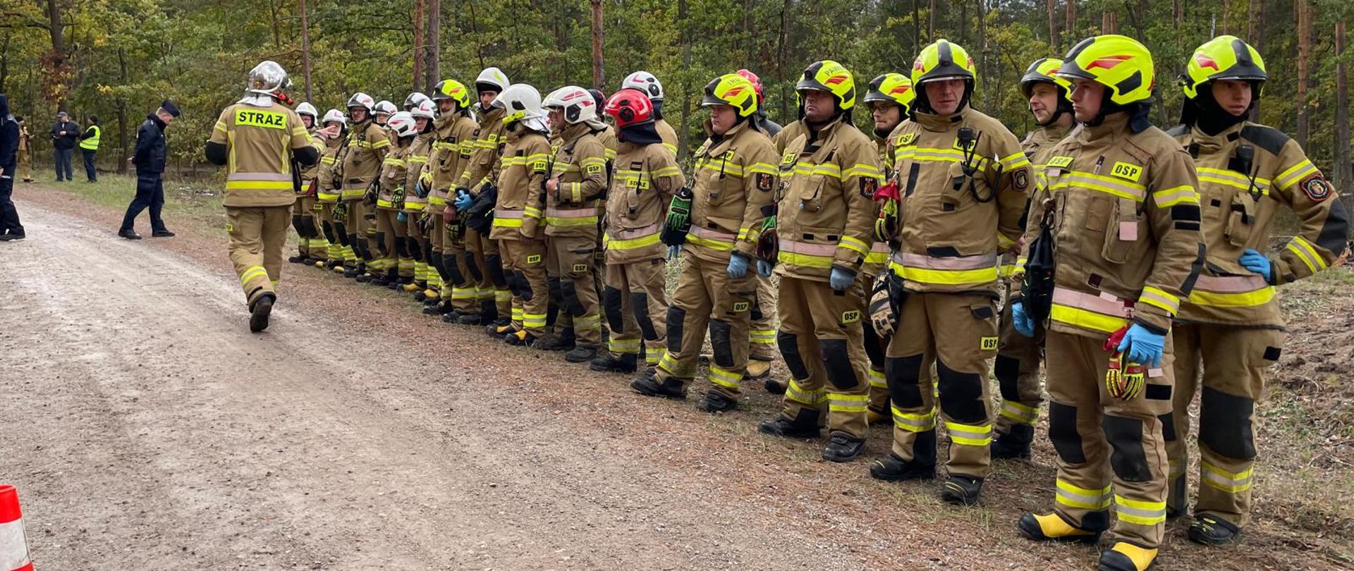 Na zdjęciu widzimy grupę strażaków stojących w szeregu na leśnej drodze gruntowej. Są ubrani w pełne umundurowanie ochronne: kaski, rękawice oraz kombinezony z odblaskowymi pasami. Jeden ze strażaków, z napisem „STRAŻ” na plecach, oddala się od grupy w lewą stronę kadru, co może sugerować, że pełni rolę dowódcy lub wykonuje konkretne zadanie.
🌲 Otoczenie to gęsto zalesiony teren, a pogoda wydaje się pochmurna — typowe warunki dla ćwiczeń terenowych lub akcji ratowniczej. W tle widać kilka innych osób, które mogą być obserwatorami, koordynatorami lub członkami innych służb.
🔥 Cała scena wygląda na dobrze zorganizowaną — może to być część szkolenia, manewrów lub przygotowań do działań w terenie. Obecność wielu strażaków i ich ustawienie wskazują na gotowość i dyscyplinę.
