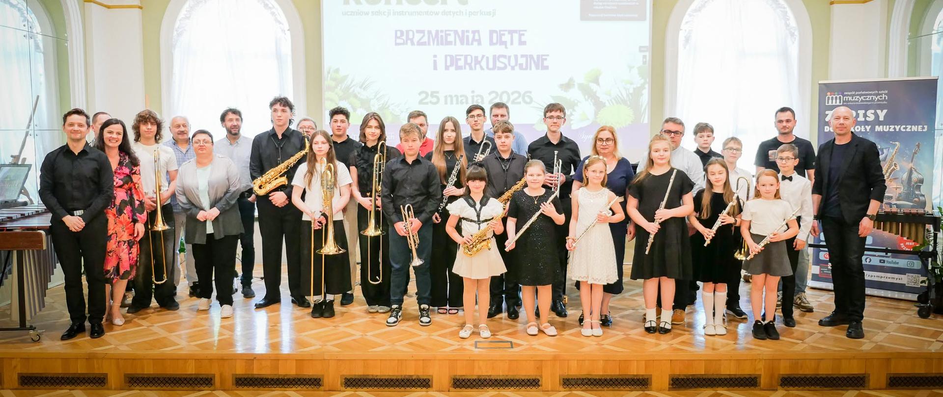 Zdjęcie przedstawia dużą grupę dzieci, młodzieży oraz dorosłych pozujących do wspólnej fotografii pamiątkowej na scenie w tej samej zabytkowej, eleganckiej sali koncertowej o pastelowo-zielonych i białych ścianach z łukowatymi oknami. Na podłodze z jasnego, błyszczącego parkietu zgromadziło się około trzydziestu osób.