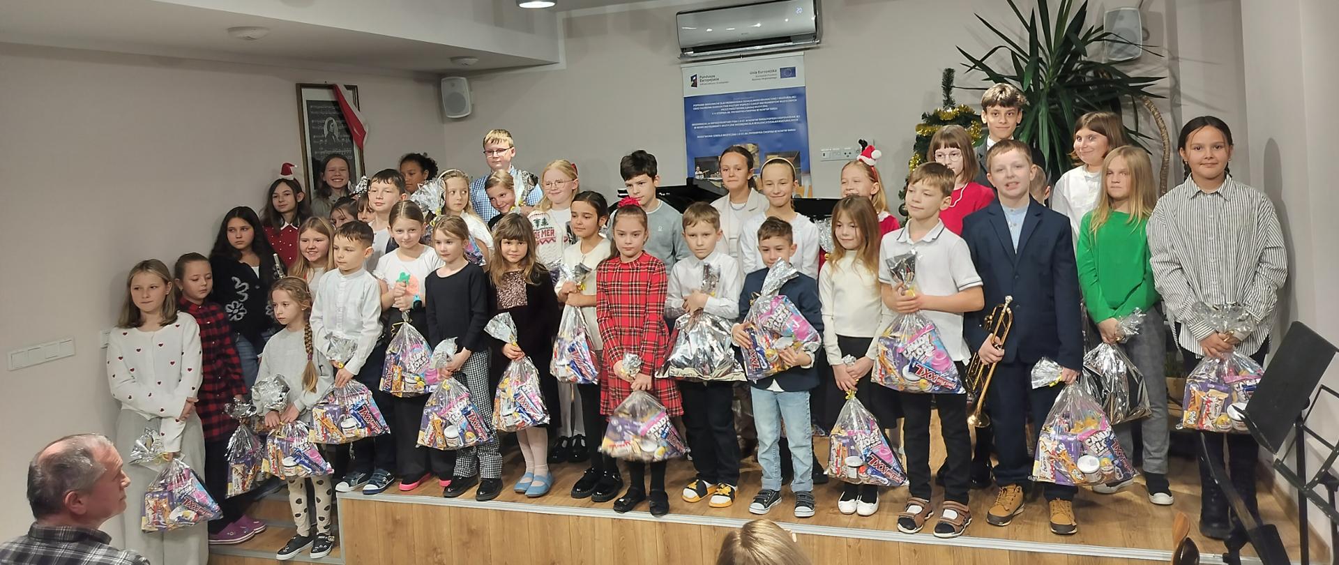 Zdjęcie przedstawia liczną grupę dzieci stojących na niskim, drewnianym podwyższeniu w jasnym pomieszczeniu. Scena wygląda na zakończenie szkolnego przedstawienia lub koncertu świątecznego. Na scenie znajduje się około 30 dzieci w różnym wieku. Większość z nich trzyma w rękach duże, foliowe torby wypełnione prezentami, słodyczami i maskotkami. Dzieci są ubrane odświętnie: Chłopcy mają na sobie koszule (białe, błękitne), a niektórzy marynarki. Jeden z chłopców po prawej stronie trzyma trąbkę. Dziewczynki ubrane są w sukienki (w tym jedna w czerwoną kratę), spódniczki lub eleganckie bluzki. Niektóre mają we włosach ozdobne opaski lub kokardy. Dzieci są ustawione w kilku rzędach, patrzą w stronę obiektywu, a na ich twarzach maluje się skupienie i zadowolenie. Scena: Niskie podest wykonany z jasnych paneli podłogowych. Tło: Ściany są jasne, o beżowym odcieniu. Na ścianie za dziećmi wisi niebieski plakat informacyjny oraz klimatyzator. Roślinność i dekoracje: Po prawej stronie, za dziećmi, widoczna jest duża zielona roślina doniczkowa (prawdopodobnie juka) oraz fragment choinki udekorowanej złotymi łańcuchami. Oświetlenie: Pomieszczenie jest mocno oświetlone przez nowoczesne, szare reflektory zamontowane na szynach pod sufitem. Widownia: Na pierwszym planie, dołem zdjęcia, widać tył głowy kobiety w różowej bluzce oraz profil mężczyzny w koszuli w kratę, którzy siedzą i obserwują dzieci. Wydarzenie odbywa się w nowoczesnej sali, prawdopodobnie w szkole muzycznej lub domu kultury, na co wskazuje obecność instrumentu i plakatu festiwalowego.