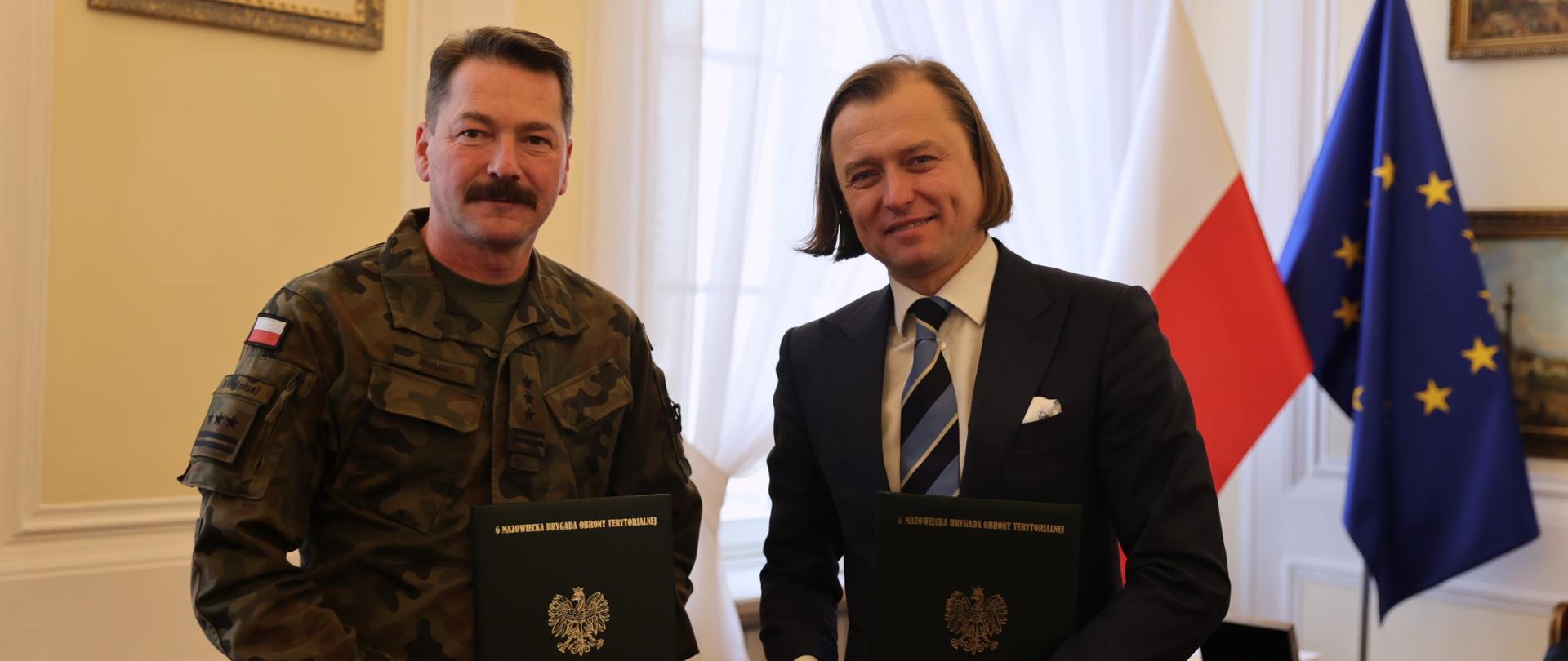 Na zdjęciu dwóch mężczyzn pozuje do zdjęcia z zielonymi teczkami 