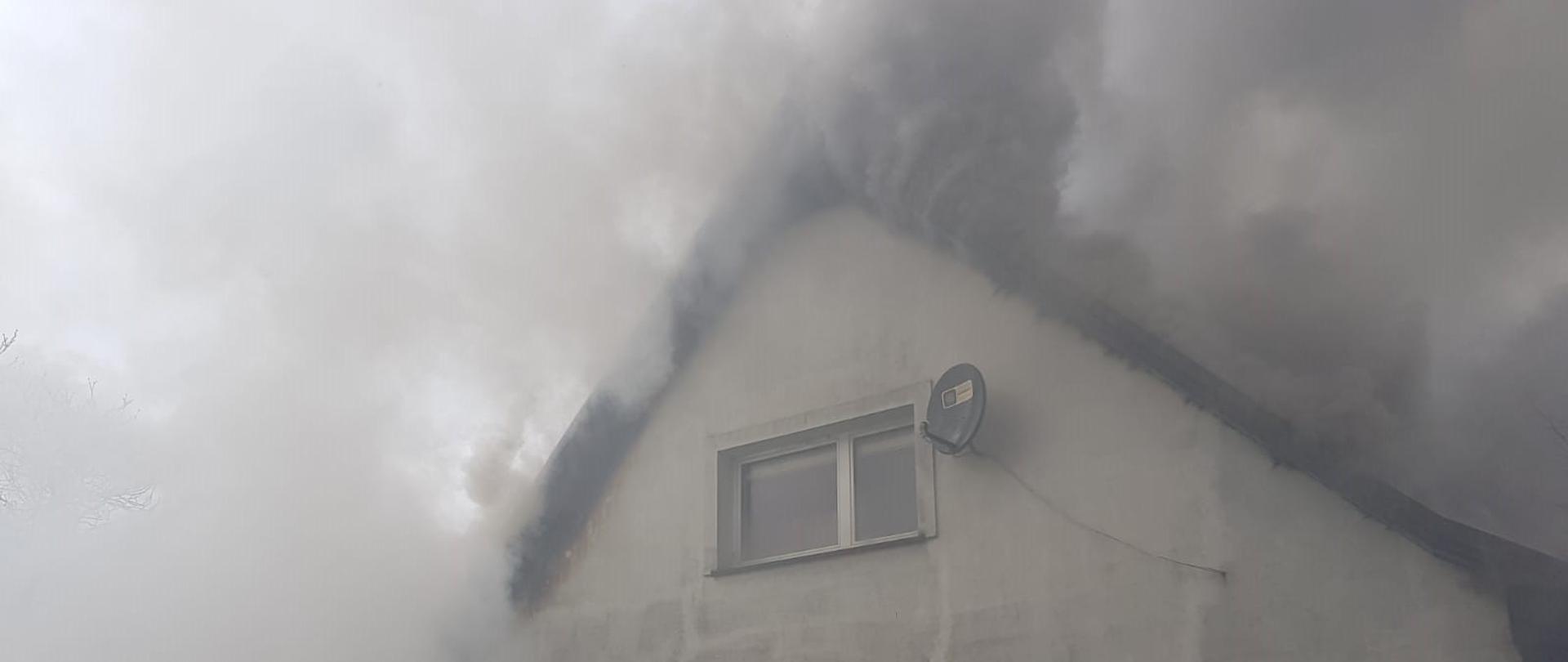 Wydobywające się kłęby szarobrunatnego dymu spod dachu. Przed budynkiem strażak z prądem gaśniczym.