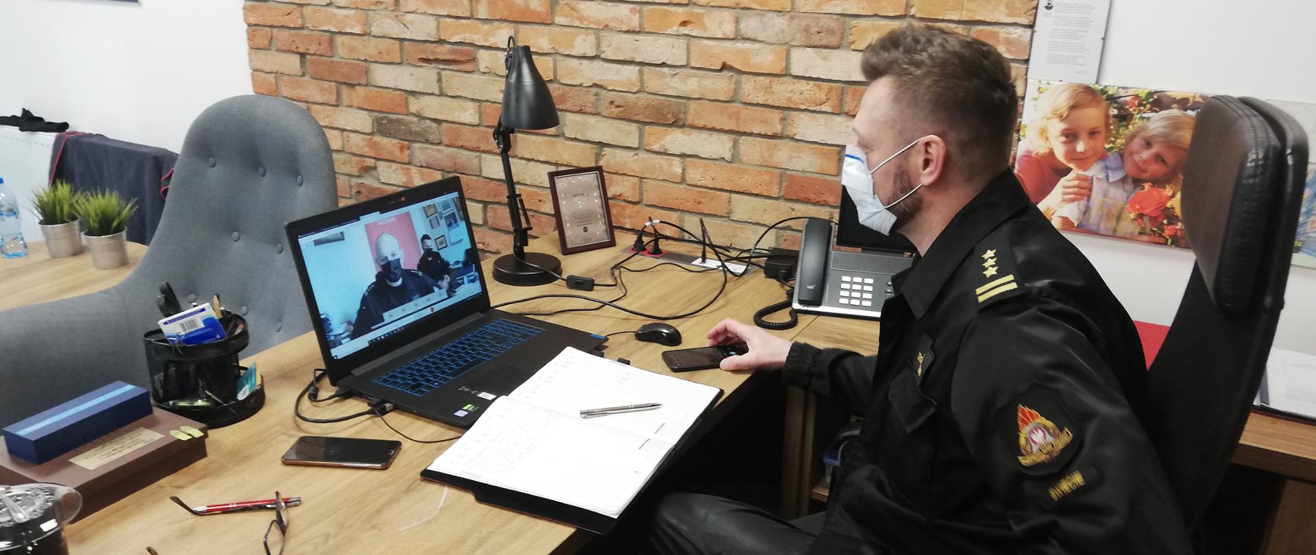 Na zdjęciu Komendant Powiatowy Państwowej Straży Pożarnej w Świeciu starszy brygadier Piotr Tkaczyński siedzi przy biurku na którym stoi laptop przez, który prowadzona jest wideokonferencja.