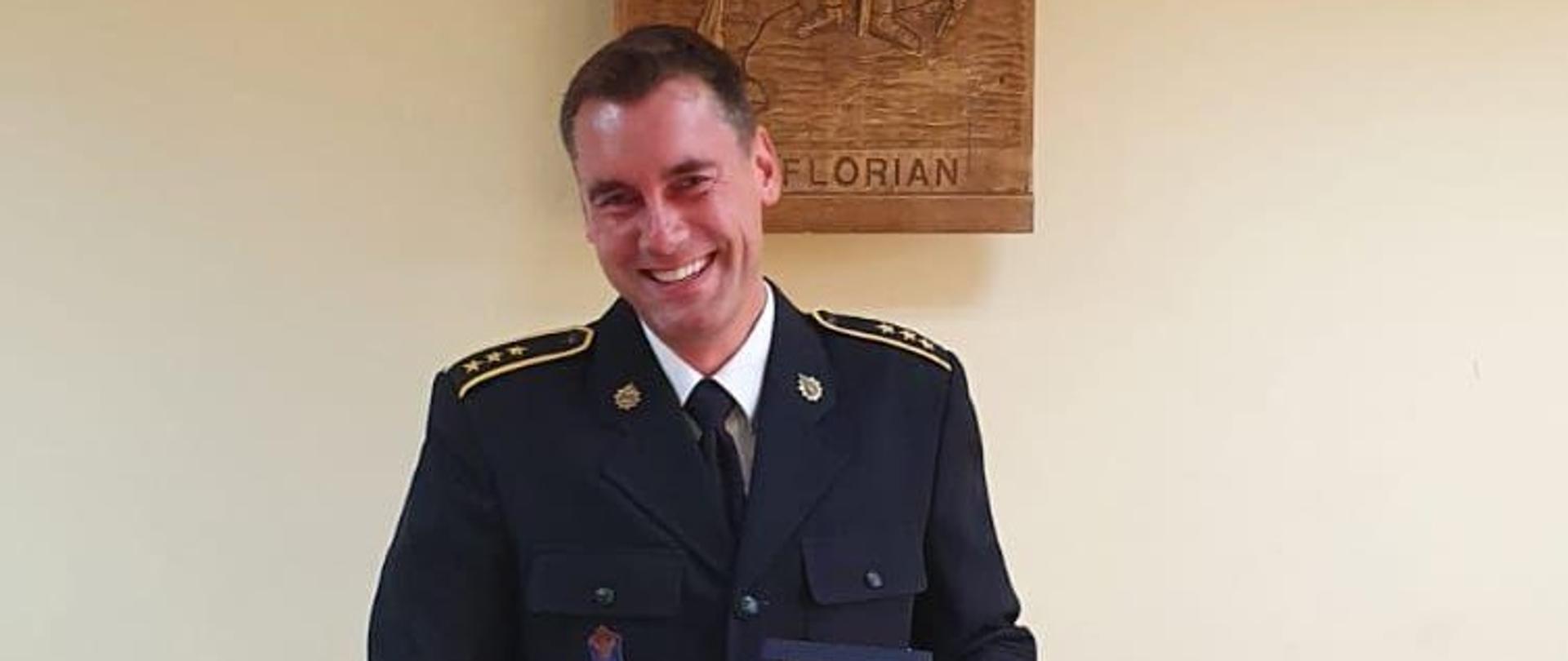 Zdjęcie przedstawia Tomasza Matuszaka - ubranego w mundur strażacki trzymającego prezent - zegarek z okazji przejścia na emeryturę 