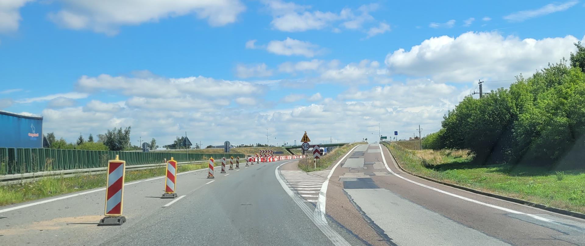 Rozjazd dróg. Lewa droga z dwoma pasami ruchu, z których lewy jest wygrodzony biało-czerwonymi pachołkami, druga - jednojezdniowa, jednokierunkowa. W oddali kolejne wygrodzenia barierami wiadukt nad drogą