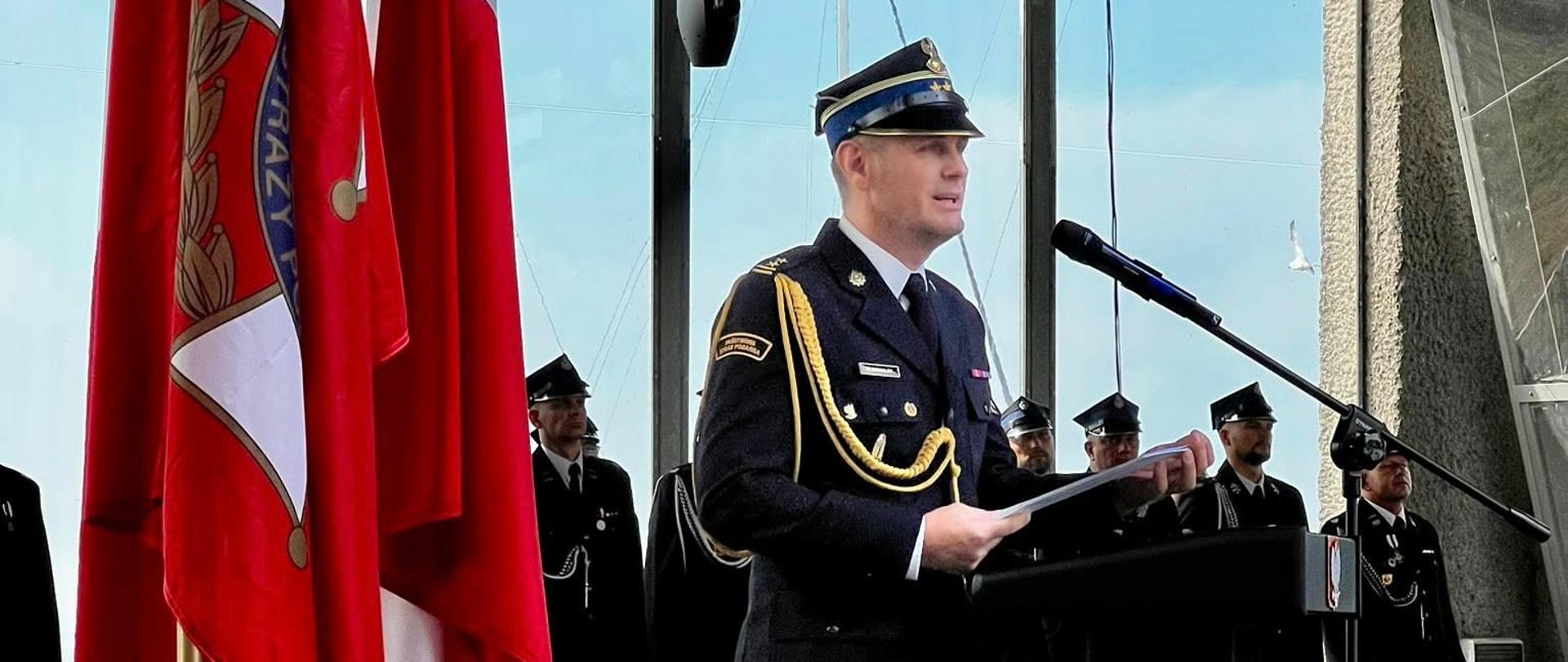 Zdjęcie przedstawia uroczystość na zewnątrz, podczas uroczystego apelu.
W centrum kadru, na podwyższonej scenie, stoi mężczyzna w galowym mundurze Państwowej Straży Pożarnej. Ma na sobie ciemny mundur z naszywkami i złotymi sznurami, a także czapkę z daszkiem. Stoi za czarnym, prostokątnym mównicą, z której wygłasza przemówienie. Przed nim znajduje się mikrofon. Mężczyzna trzyma w dłoniach dokumenty.
Po jego prawej stronie, w tle, widać grupę innych mężczyzn w podobnych mundurach. Stoją w szyku, zwróceni w kierunku mównicy. Po lewej stronie kadru znajdują się cztery flagi na masztach. Dwie z nich to flagi Polski, a dwie to sztandary strażackie z godłem Państwowej Straży Pożarnej.
W tle widoczna jest scena lub konstrukcja z metalowych kratownic. Pogoda wydaje się być słoneczna, a na niebie widać lekkie chmury
