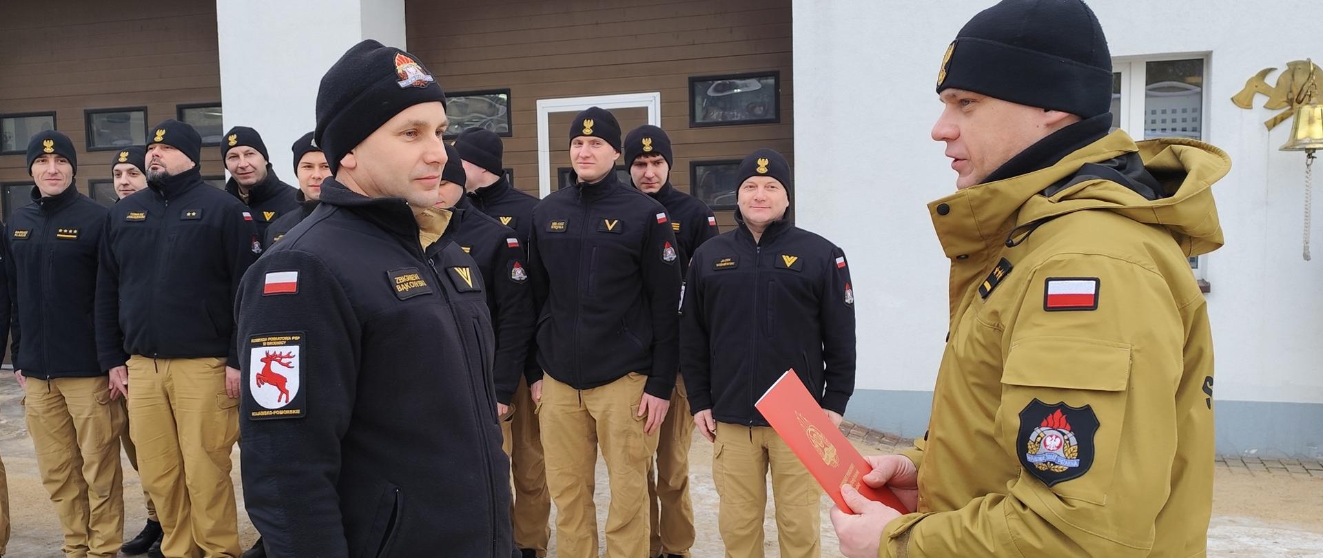 Zdjęcie przedstawia Komendanta Powiatowego PSP w Brodnicy - st. kpt. mgr Krzysztofa Natuckiego wręczającego list gratulacyjny z okazji jubileuszu 30 lat służby i pracy st. ogn. Zbigniewowi Bąkowskiemu. Za nimi stoją w dwuszeregu funkcjonariusze Komedny w mundurach służbowych.