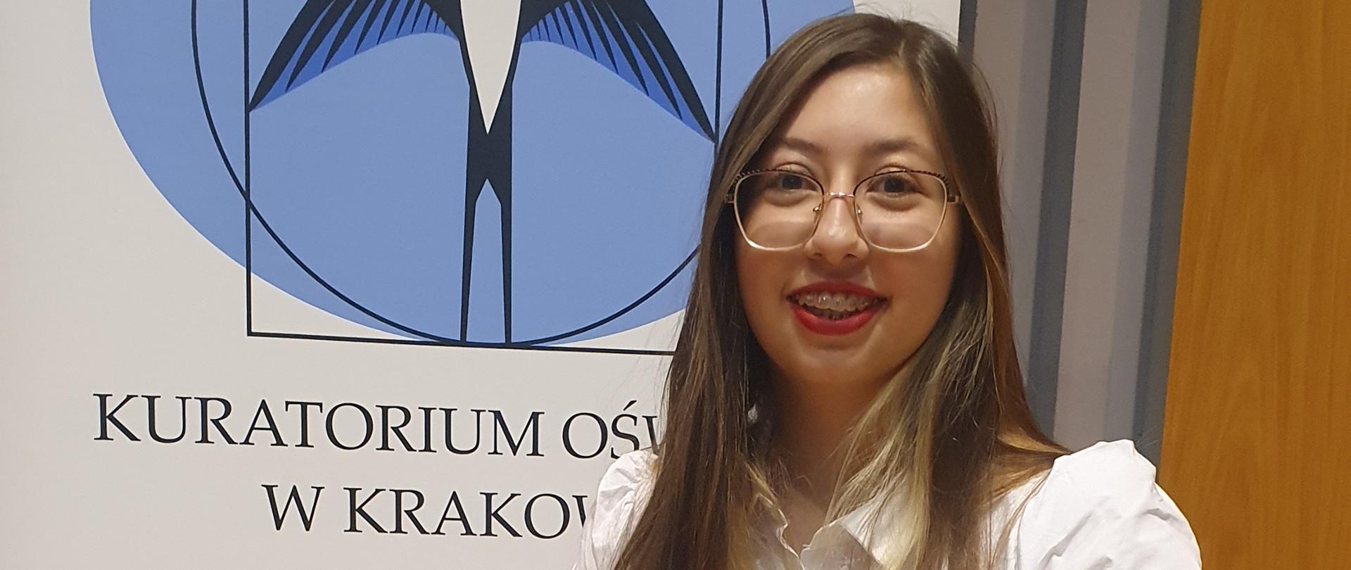 Na zdjęciu Katarzyna Kurzydło uczennica kl. 4C Państwowego Liceum Sztuk Plastycznych im. Antoniego Kenara w Zakopanem. Postać ubrana jest w białą sukienkę. Katarzyna ma rozpuszczone długie włosy, jest uśmiechnięta. Uczennica ma założone okulary ze złotymi oprawkami. Dziewczyna trzyma w rękach dyplom gratulacyjny od Prezesa Rady Ministrów Mateusza Morawieckiego. Na drugim planie znajduje się stojący baner z napisem Kuratorium Oświaty w Krakowie. W górnej części baneru znajduje się logo Kuratorium - jaskółka na błękitnym kole wpisanym w kwadrat. Na dole baneru znajduje się szary element wycięty z koła.