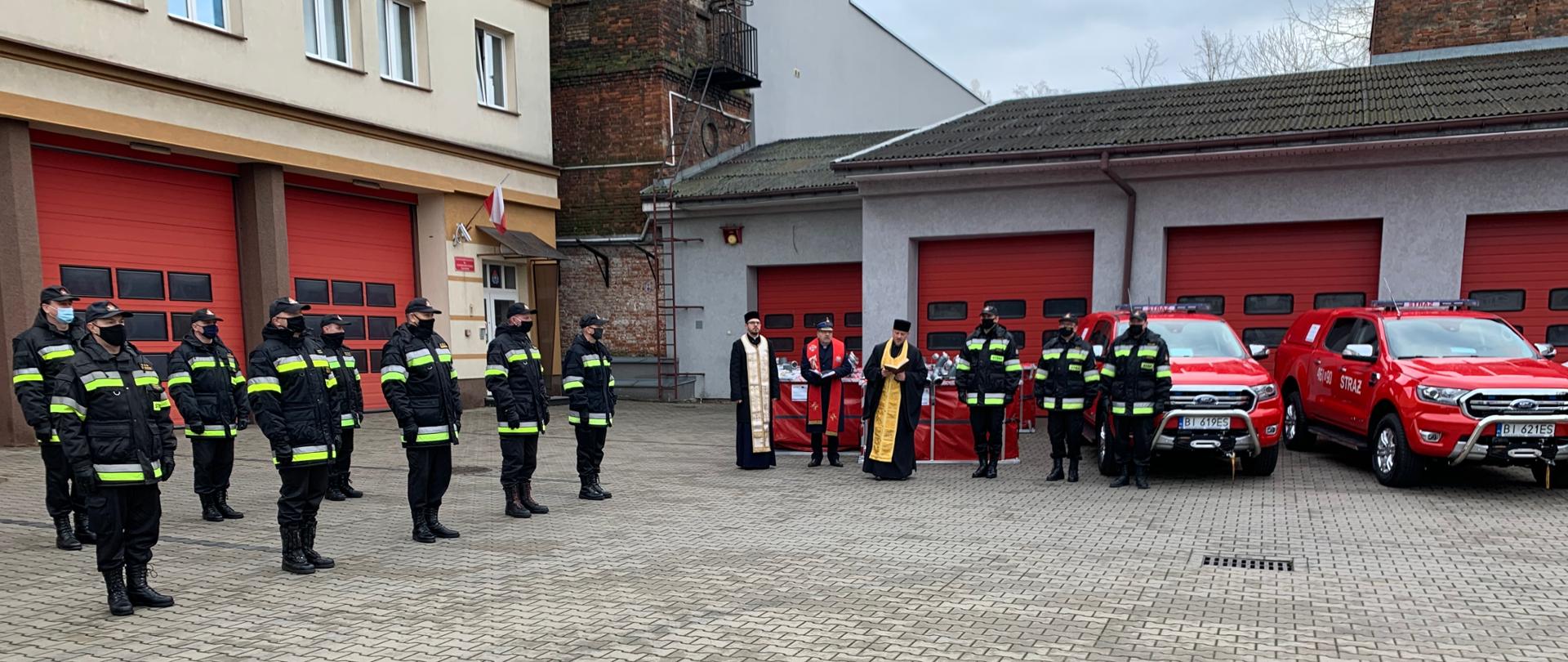 Zdjęcie obrazujące strażaków strażaków ustawionych w dwuszeregu w umundurowaniu dowódczo-sztabowym i kapelani strażaccy w szatach liturgicznych ustawieni podczas uroczystego przekazania sprzętu i pojazdów zakupionych w ramach Programu Współpracy Transgranicznej Polska-Rosja 2014-2020 