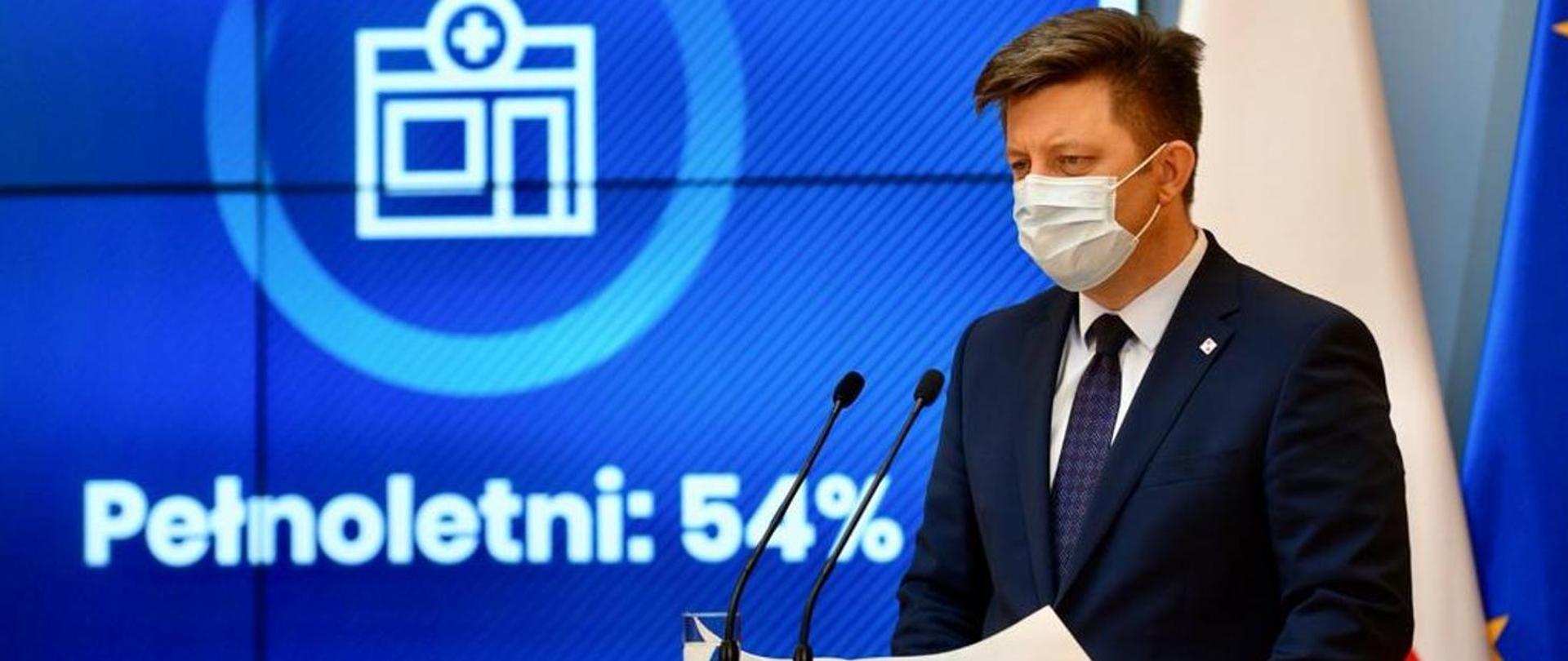 Po prawej stronie mężczyzna w maseczce, w garniturze, przed dwoma mikrofonami, na tle niebieskiego napisu pełnoletni 54%