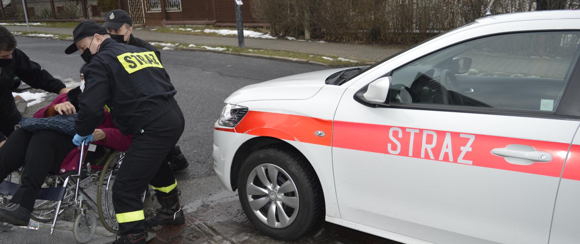 Na zdjęciu znajdują się druhowie Ochotniczej Straży Pożarnej, którzy pomagają w wprowadzeniu osoby na wózku do samochodu. Po prawej stronie częściowo widoczny jest również samochód Państwowej Straży Pożarnej.