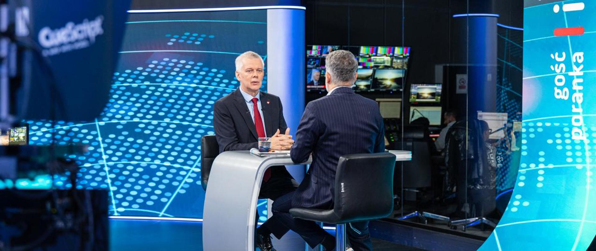 Na zdjęciu wnętrze studia telewizyjnego. Przy szarym stole na czarnych krzesłach siedzą: minister Tomasz Siemoniak (po lewej) oraz redaktor Mariusz Piekarski (po prawej, tyłem do obiektywu). Na stole szklanka z wodą i gazety. W tle przez szybę widać wnętrze redakcji - dziennikarzy, monitory i inny sprzęt. Po prawej stronie zdjęcia biały napis na niebieskim tle "Gość Poranka". Za ministrem widać ściankę wizyjną z biało-niebieskimi elementami graficznymi. Po lewej stronie zdjęcia widać fragment czarnej kamery.