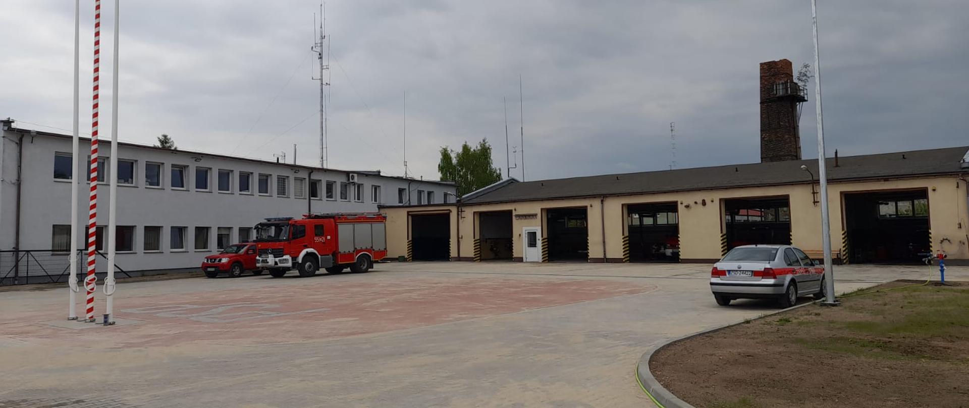 Zdjęcie przedstawia plac wewnętrzny KP PSP Świdwin, w tle znajdują się maszty flagowe, budynek komendy, bramy garażowe oraz samochód strażacki.