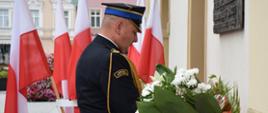 Rawicki rynek. Strażak w umundurowaniu wyjściowym składa wiązanke biało-czerwonych kwaitów pd tablicą pamiątkową umieszczona na budynku ratusza.. W tle stojak z flagami biało-czerwonymi. 