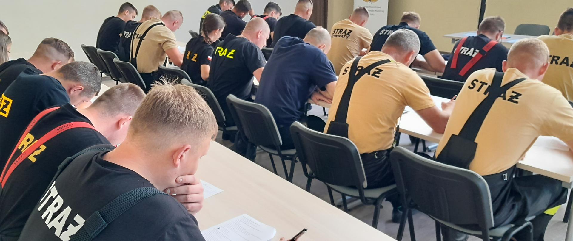 Grupa osób w jasnych lub ciemnych koszulkach z napisem straż siedzi w ławkach. Osoby znajdują się tyłem do aparatu z którego zrobiono zdjęcie.