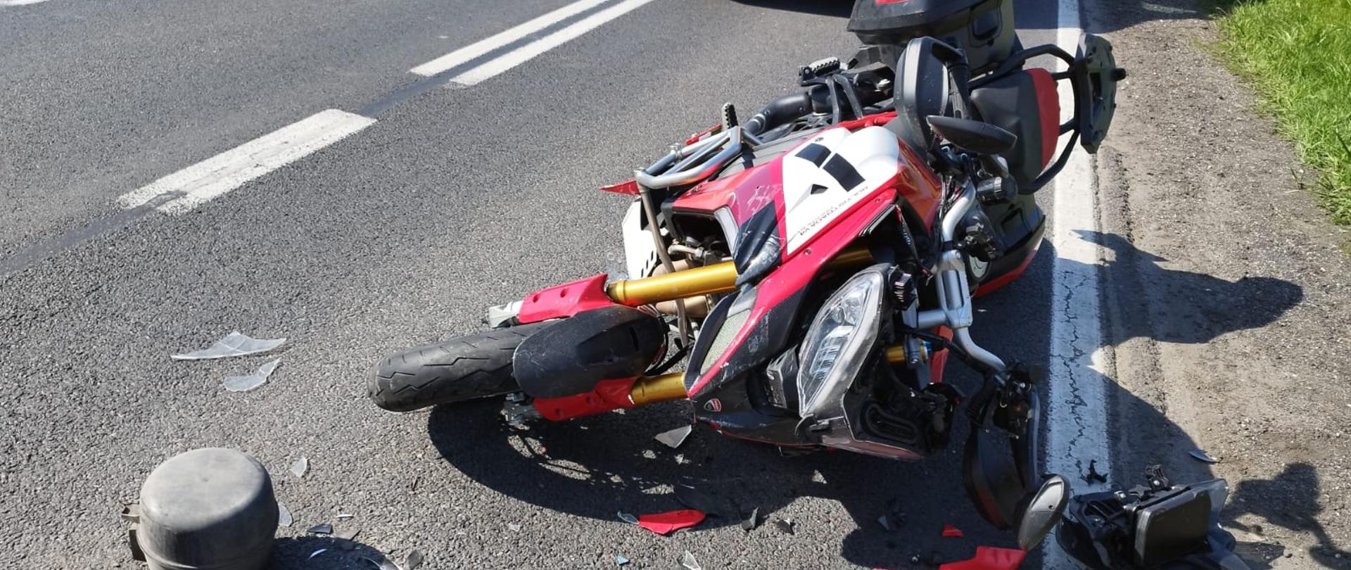 Motocykl leży na drodze po zderzeniu z samochodem dostawczym. W tle stoi karetka pogotowia